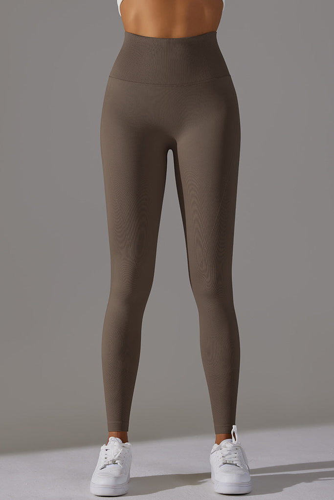 Kvinde iført mørkebrune leggings og top, let drejet med vægten i det ene ben.

